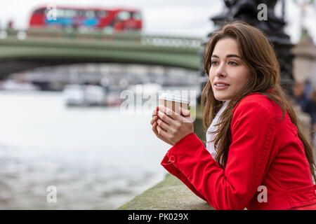 Ragazza o giovane donna in un cappotto rosso di bere il caffè in una tazza monouso accanto a Westminster Bridge con il bus rosso a due piani in background, Londra, Foto Stock