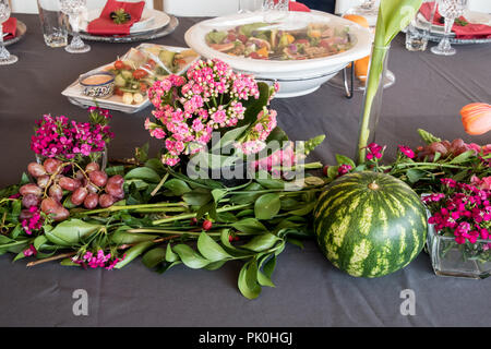 Un tavolo pranzo con display floreali di bellissimi fiori arancione con colorati di rosa e regime rossastro in primo piano con un giro di cocomero e uva Foto Stock