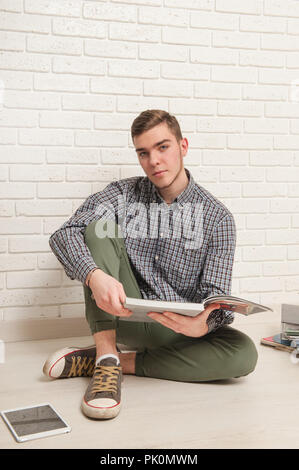 Giovane uomo si siede con i libri di testo sul pavimento contro un muro di mattoni in background Foto Stock