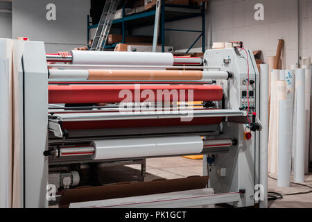 Grande stampa professionale laminatore, armati con rossi grandi rotoli di carta lucida e piccoli tubi di pellicola trasparente. Foto Stock