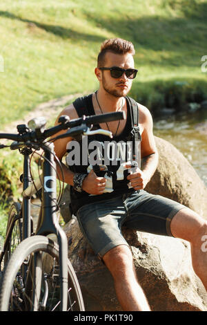 Elegante giovane con una bici seduto su di una roccia. T-shirt nera con stampa 23 Foto Stock