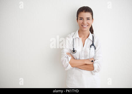 Gentile felice femmina giovane infermiere o medico con uno stetoscopio attorno al collo in piedi con le braccia incrociate contro un muro bianco con spazio copia sorridente Foto Stock