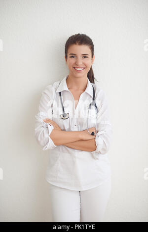 Gentile felice femmina giovane infermiere o medico con uno stetoscopio attorno al collo in piedi con le braccia incrociate contro un muro bianco con spazio copia sorridente Foto Stock