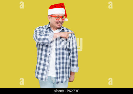 Positivo uomo di mezza età in cappuccio rosso, occhiali e camicia a scacchi in piedi e puntare il dito per svuotare lo spazio di copia e toothy sorridente, guardando a ca Foto Stock