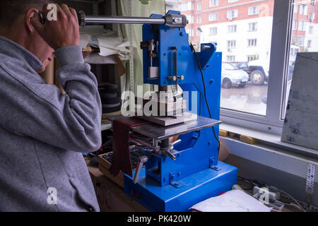 L'uomo in officina preme la leva sulla pressa per ottenere il logo in rilievo e lettere sul manufatto in pelle che riscalda il cliché e s Foto Stock