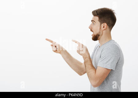 Studio shot di sorpreso e impressionato bello guy standing in profilo con ganascia interrotte e sguardo incuriosito di puntamento e guardando verso sinistra attoniti e senza parole su sfondo bianco Foto Stock