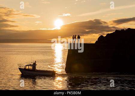 Silhouette di persone che saltano dalla parete del porto al tramonto nella luce del sole di setting come una barca che passa, Portstewart, Co. Derry, Irlanda del Nord Foto Stock