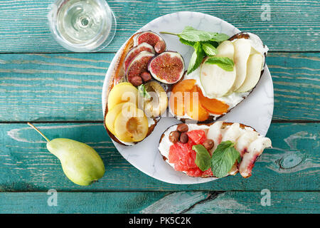 Spuntini di frutta per il vino. Piastra con diversi tipi di frutta bruschetta verde sul tavolo di legno con un bicchiere di vino bianco, vista dall'alto Foto Stock