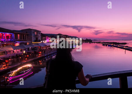 Donna turistica, con ritorno alla macchina fotografica, guardando il tramonto sul ponte del porto a Lefkada (Lefkas), Grecia Foto Stock