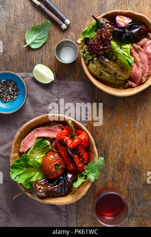 Bistecca alla griglia con verdure alla griglia servita nella piastra di pane sul tavolo di legno con un bicchiere di vino rosso, vista dall'alto Foto Stock