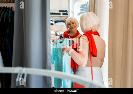 Guardando bene dai capelli grigi donna guardando lo specchio nella sala di montaggio del negozio di abbigliamento Foto Stock