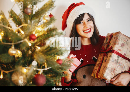 Felice giovane donna in santa hat holding pila di confezioni regalo al golden bellissimo albero di natale con le luci in sala festosa. felice vacanza invernale atmosph Foto Stock