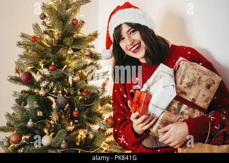 Felice ragazza in santa hat holding molte confezioni regalo al golden bellissimo albero di natale con le luci in sala festosa. felice vacanza invernale momen atmosferica Foto Stock