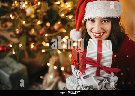 Felice ragazza ritratto in santa hat holding molte confezioni regalo al golden bellissimo albero di natale con le luci in sala festosa. vacanze invernali mo atmosferica Foto Stock