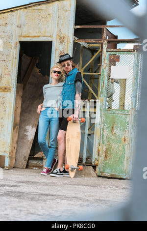 Ragazzo con tatuaggi e elegante ragazza in posa con longboard e guardando lontano Foto Stock