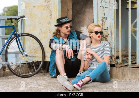 Ragazzo con tatuaggi e elegante ragazza seduta vicino alla parete e bicicletta Foto Stock