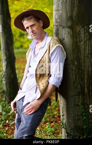 Un uomo vestito come un cowboy si appoggia contro un albero di fumare una sigaretta Foto Stock