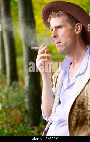 Un uomo vestito come un cowboy si appoggia contro un albero di fumare una sigaretta Foto Stock