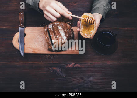 Vista superiore mani femminili fanno un sandwich di pane di segale e miele scuro sul tavolo di legno accanto alla tazza di tè nero o caffè. Composizione di semplici piatti rustici. Foto Stock