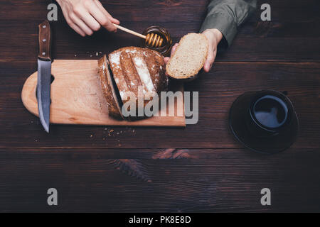 Vista superiore mani femminili dip cucchiaio di miele in miele per rendere dolce il pane di segale sandwich scuro sul tavolo di legno accanto alla tazza di tè nero o caffè. Composizione Foto Stock