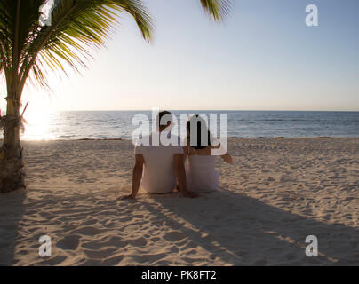 Carino coppia guardando il tramonto su una spiaggia con palme. Retro del giovane seduta nella sabbia su una spiaggia durante una vacanza tropicale. Foto Stock