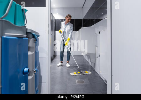 La signora di pulizia con straccio il pavimento in mens restroom Foto Stock