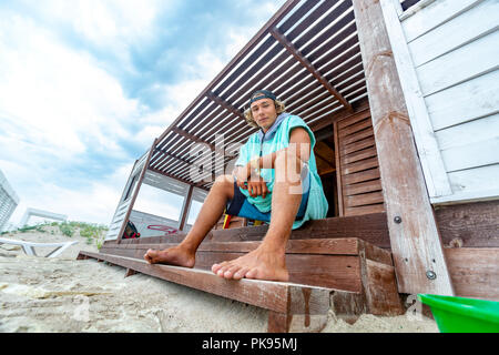 Il surfer si siede sulla terrazza sulla spiaggia guardando in lontananza. Foto Stock