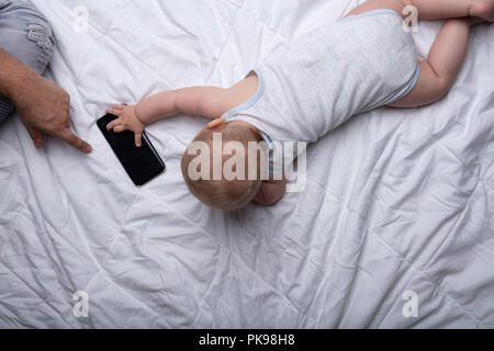Piccolo neonato per raggiungere un telefono cellulare toccando lo schermo in un gesto tattile con la mano di suo padre amorevole vicino su un letto bianco fr Foto Stock