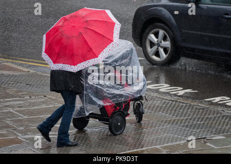 Regno Unito Meteo. Una donna con un ombrello rosso spingendo il suo bambino in una carrozzina mentre catturati in una pesante pioggia. Foto Stock
