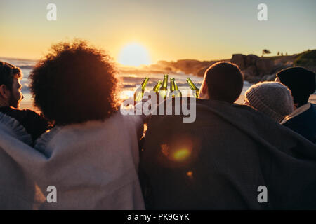 Gruppo di amici cheers con birre alla spina presso la spiaggia. I giovani seduti insieme in spiaggia durante il tramonto e avente una parte. Foto Stock