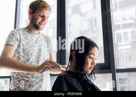 Giovane bella donna con i capelli tagliati dal parrucchiere. Giovane maschio parrucchiere sorridente e rendendo l'acconciatura per il cliente. Foto Stock