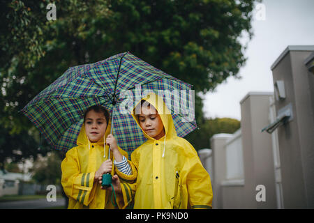 Due bambine con ombrello aperto sul giorno di pioggia. Gemelle indossando vestiti impermeabili a camminare insieme sulla strada. Foto Stock