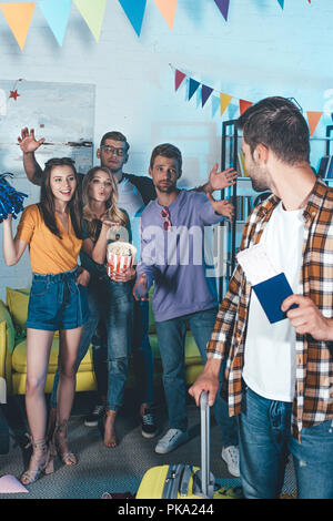 Giovane uomo con passaporto e valigia guardando amici alla festa di commiato Foto Stock