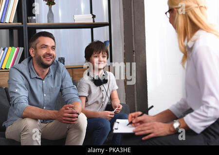 Vista laterale del consigliere femmina iscritto nella clipboard mentre il padre e il figlio con le cuffie seduti sulla sessione di terapia in office Foto Stock