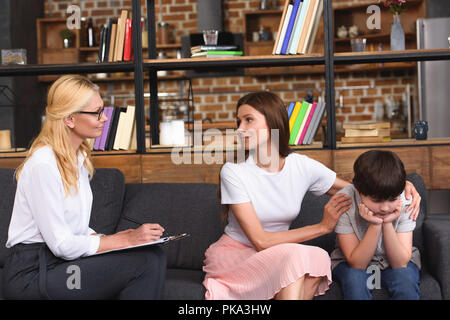 Vista laterale del consigliere femmina parlando a madre mentre lei ha sottolineato che abbraccia il figlio sulla sessione di terapia Foto Stock