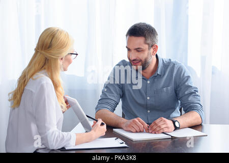 Vista laterale del consigliere femmina puntando sulla tavoletta digitale schermo con la penna per sorridere paziente di sesso maschile a tavola in office Foto Stock
