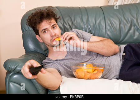 Uomo con telecomando e patatine guardando la TV a casa Foto Stock