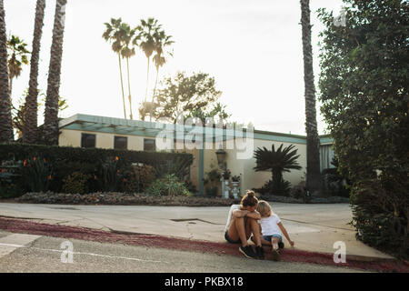 Due giovani ragazze a sedersi su un cordolo di strada al di fuori di una casa; Los Angeles, California, Stati Uniti d'America Foto Stock