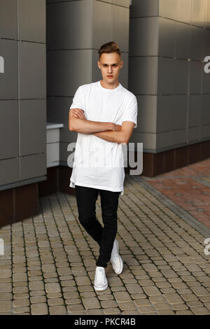 Bel giovane hipster elegante Uomo in camicia bianca e pantaloni neri e scarpe di colore bianco in posa vicino al moderno muro grigio. Foto Stock