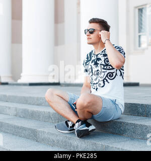 Giovane uomo elegante con occhiali da sole seduti sulle scale e ascolto di musica. Foto Stock