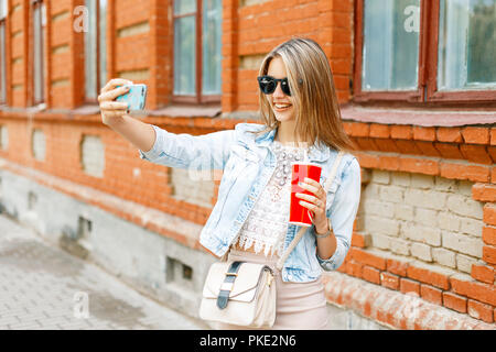 Bella donna allegra con un sorriso in una camicia di denim azienda red cup con drink e fotografati stessi sul telefono su un giorno d'estate. Foto Stock