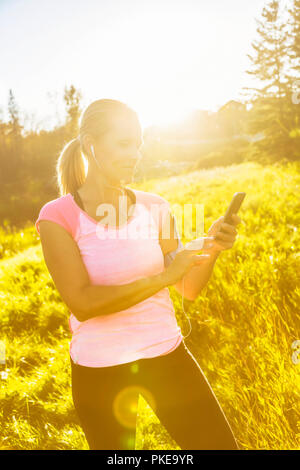 Una donna attraente di mezza età che indossa abbigliamento attivo si ferma per SMS mentre fuori correre accanto a un torrente in un parco della città in una calda... Foto Stock