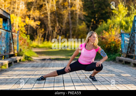 Un attraente donna di mezza età delle pause per tendere le sue gambe su un ponte durante una corsa su un bellissimo autunno caldo la sera in un parco della città Foto Stock