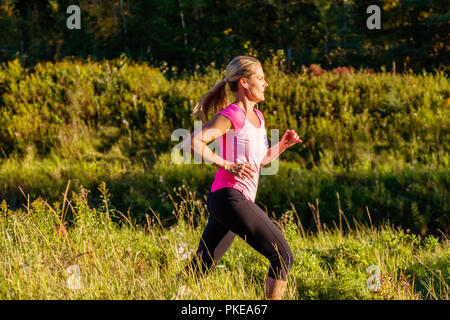 Un attraente donna di mezza età che indossa active wear e ascolto di musica corre lungo un ruscello in un parco della città al tramonto in una calda serata d'autunno Foto Stock