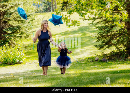 Una madre a piedi attraverso un parco su una calda giornata di caduta con la sua bambina che indossa un abito di partito e sono entrambe le portanti a forma di stella palloncini Foto Stock