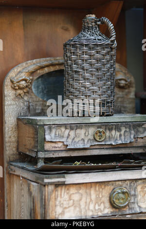 Tanica in cesto di vimini su un tavolo di legno. Vecchio demijohn avvolto in vimini bottiglia di vetro sul vecchio tavolo in legno Foto Stock