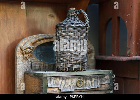 Tanica in cesto di vimini su un tavolo di legno. Vecchio demijohn avvolto in vimini bottiglia di vetro sul vecchio tavolo in legno Foto Stock