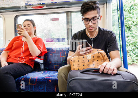 Londra Inghilterra,Regno Unito,Piccadilly Circus Line,treno interno uomo donna maschio, seduta con smartphone auricolari bagagli passeggeri Foto Stock