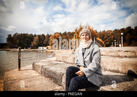 Outdoor Lifestyle atmosferica foto del giovane bella darkhaired donna in maglia hat, in un cappotto grigio e pantaloni neri si siede sul molo accanto a Foto Stock