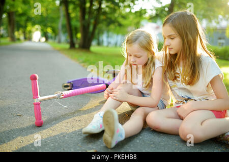 Adorabile ragazza consolante la sua sorellina dopo che lei è caduto il suo scooter a summer park. Bambino di farsi male mentre cavalcate un kick scooter. Famil attivo Foto Stock